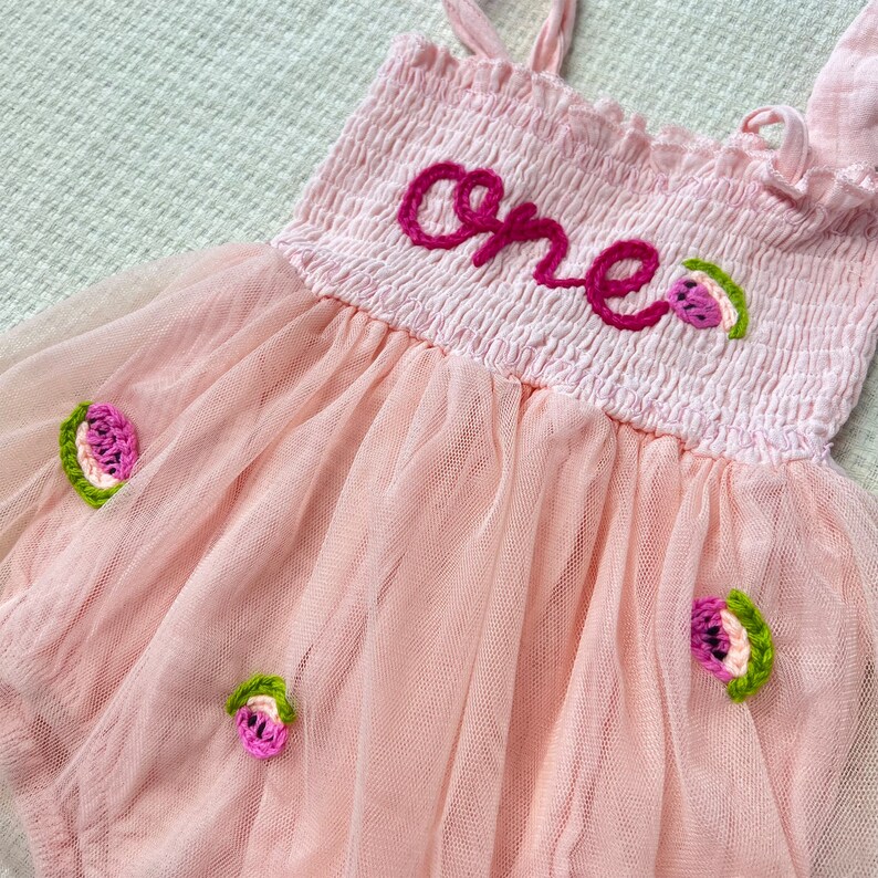 May include: Pink tulle baby dress with embroidered watermelon details and the word "one" in pink thread.