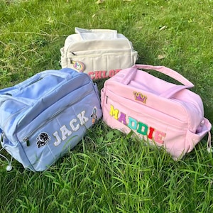 May include: Three personalized baby bags in pastel colors sit on green grass. A light blue bag has the name "JACK" embroidered on it, a pink bag says "MADDIE", and a cream-colored bag says "CHLOE". Each bag has a unique patch.