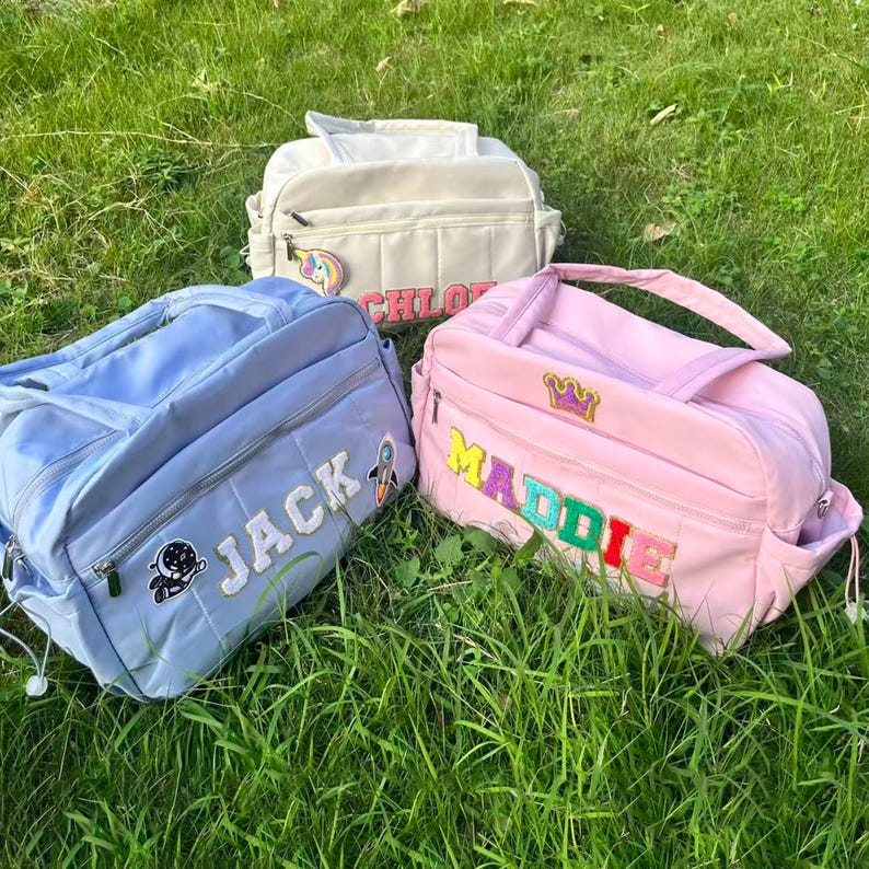 May include: Three personalized baby bags in pastel colors sit on green grass. A light blue bag has the name "JACK" embroidered on it, a pink bag says "MADDIE", and a cream-colored bag says "CHLOE". Each bag has a unique patch.