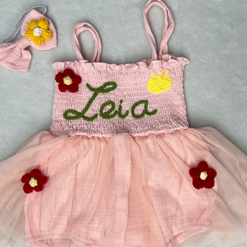 May include: Pink tulle baby dress with a crocheted bodice featuring the name "Leia" in green yarn, a yellow crown, and red flowers. The dress comes with a matching headband with a yellow flower.