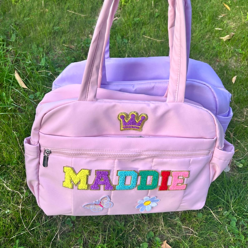 May include: Pink quilted tote bag with the name "MADDIE" in colorful letters. Features a gold crown patch and a butterfly and flower applique. A lavender bag is visible behind it.