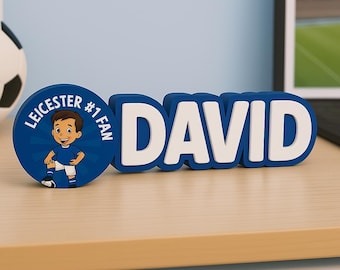 Puede incluir: Un letrero con nombre personalizado azul y blanco que dice "DAVID" en letras en negrita. Un emblema circular azul con el texto "LEICESTER #1 FAN" y un jugador de fútbol de dibujos animados está al lado del letrero con el nombre. Los artículos están sobre una superficie marrón claro.