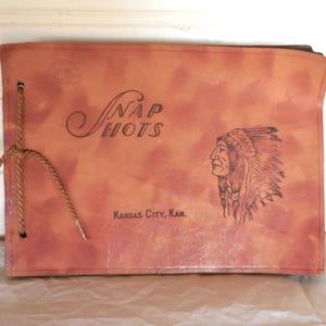 May include: Vintage photo album with the word "Snapshots" in elegant script. The cover is a reddish-brown color and features an illustration of a Native American chief and the text "Kansas City, Kan." A twine cord binds the album.