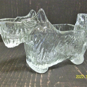 May include: A clear glass figurine in the shape of a Scottish Terrier dog. The dog has a textured surface and is posed in a sitting position. The figurine has a rectangular opening on its back, suggesting it could be a container.