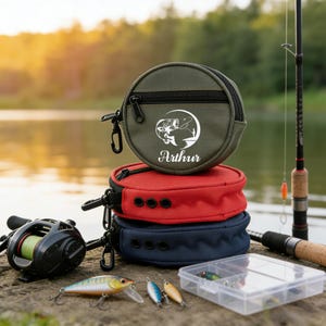May include: Three fishing tackle pouches in olive green, red, and navy blue are stacked. The top pouch has a zipper and the name "Arthur" printed below a fishing graphic. Fishing lures and a fishing rod are also visible.