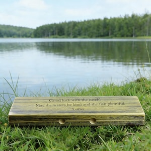 Könnte beinhalten: Eine hölzerne Angelkiste mit dem Text "Good luck with the catch! May the waters be kind and the fish plentiful. Lucas." Die Kiste ist hellgrün und braun, mit zwei kreisförmigen Löchern.