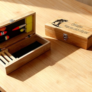 May include: A bamboo fishing tackle box, open to reveal compartments and floats. The closed box features a fish graphic and the text "Christopher Hope you catch a big one!" A small metal clasp secures the lid. The box is on a wooden surface.