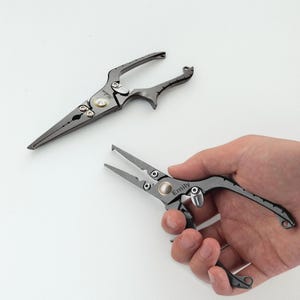 May include: Two pairs of dark gray fishing pliers. One pair is open, and the other is being held in a hand. The pliers have a pointed tip and a curved handle. The pliers have the name "Emily" engraved on them.