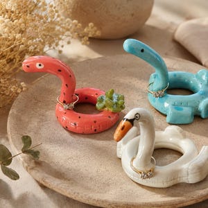 May include: Three ceramic ring holders shaped like pool floats. One is a coral pink snake with a green succulent, one is a light blue dinosaur, and one is a white swan with an orange beak. Each float has a ring resting on its neck.