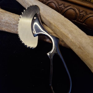 Könnte beinhalten: Eine chirurgische Knochensäge aus silberfarbenem Metall mit einer halbkreisförmigen, gezahnten Klinge, die an einem schlanken, länglichen Griff mit einer Schlaufe am Ende befestigt ist. Das Werkzeug liegt an einem hellen, strukturierten Knochenfragment.