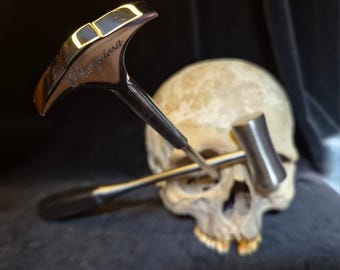 May include: A polished silver-toned metal tool with a T-shaped head featuring the engraved text "Furiosa" and a second smaller cylindrical metal hammer tool, both positioned in front of a realistic human skull with visible teeth and eye sockets.