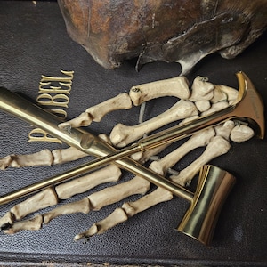 Op de afbeelding: Een verzameling messing rookpijpen en een botten handsteun op een donker, getextureerd oppervlak. De pijpen hebben een gepolijste afwerking en de botten hand is lichtbeige van kleur. Het woord "BIJBEL" is zichtbaar in gouden letters.