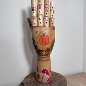 May include: Wooden hand sculpture with a fall theme. The fingers spell out "FALL" with leaf illustrations. The palm features a pumpkin and floral design. The base has red and white mushroom illustrations. The sculpture sits on a wooden round base.