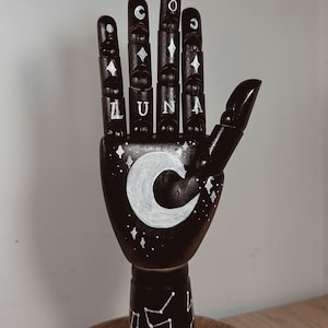 Puede incluir: Escultura de mano de madera negra con diseños celestiales blancos. La palma presenta una luna creciente blanca y la palabra "LUNA". Los dedos tienen diamantes blancos y acentos de luna creciente. La base tiene un diseño de constelación.