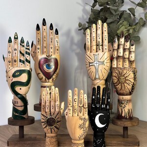 Può includere: Mani articolate in legno con vari disegni dipinti. Una mano presenta un serpente verde, un'altra un cuore con un occhio, e altre hanno motivi di sole, luna e croce. Alcune dita hanno unghie dipinte e testo.