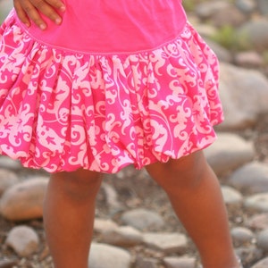 K&ouml;nnte beinhalten: Ein junges M&auml;dchen tr&auml;gt ein wei&szlig;es Shirt und einen rosa Rock mit wei&szlig;em Blumenmuster. Der Rock ist in der Taille gerafft und hat einen Ballonrock-Saum. Das M&auml;dchen steht auf einer felsigen Oberfl&auml;che.