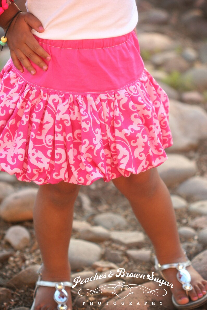K&ouml;nnte beinhalten: Ein junges M&auml;dchen tr&auml;gt ein wei&szlig;es Shirt und einen rosa Rock mit wei&szlig;em Blumenmuster. Der Rock ist in der Taille gerafft und hat einen Ballonrock-Saum. Das M&auml;dchen steht auf einer felsigen Oberfl&auml;che.