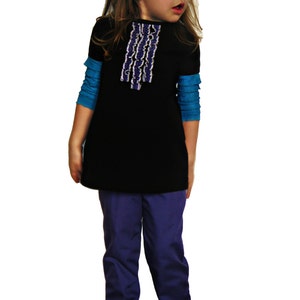 Puede incluir: Una ni&ntilde;a peque&ntilde;a viste una t&uacute;nica negra con un dise&ntilde;o de volantes blancos y morados, sobre una camiseta azul de manga larga. Tambi&eacute;n lleva pantalones morados y zapatos Mary Jane negros. Un lazo negro adorna su cabello.