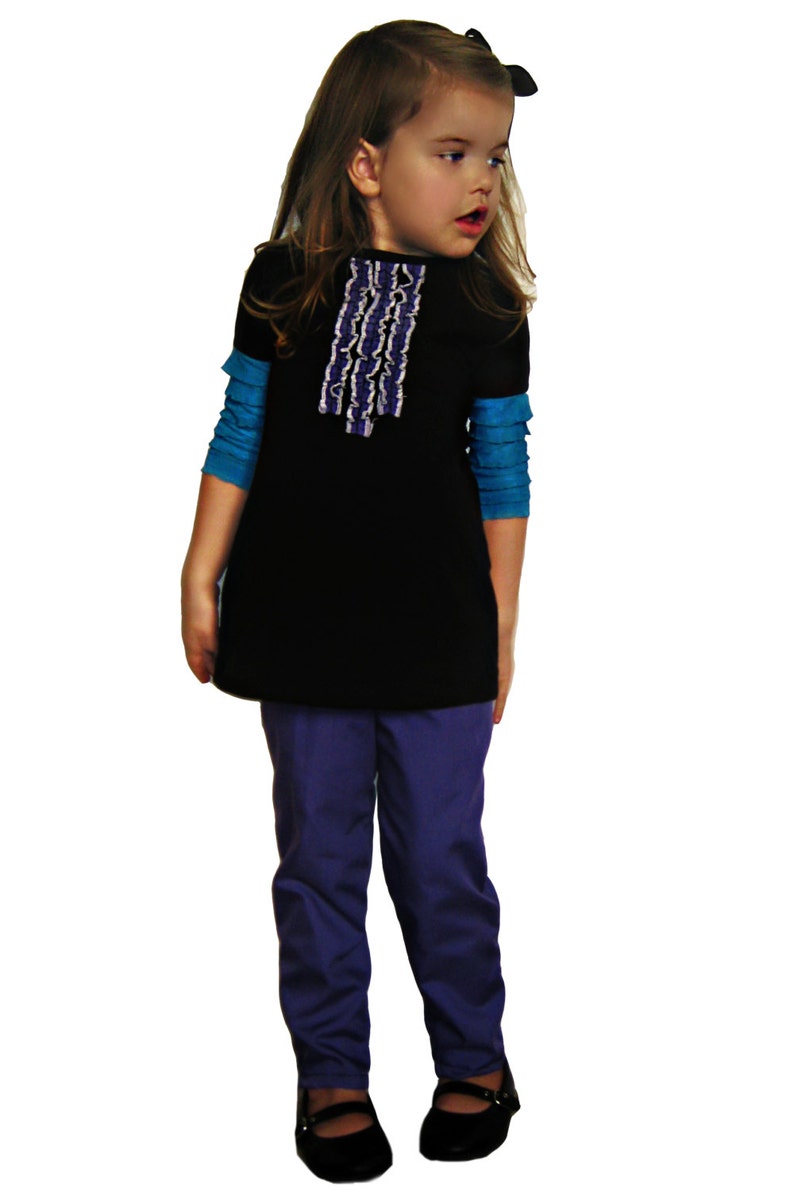 Puede incluir: Una ni&ntilde;a peque&ntilde;a viste una t&uacute;nica negra con un dise&ntilde;o de volantes blancos y morados, sobre una camiseta azul de manga larga. Tambi&eacute;n lleva pantalones morados y zapatos Mary Jane negros. Un lazo negro adorna su cabello.