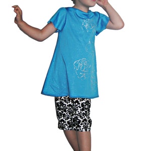 Puede incluir: Una ni&ntilde;a peque&ntilde;a con un vestido turquesa con detalles de rosas plateadas y pantalones capri con estampado en blanco y negro. Tambi&eacute;n lleva una diadema a juego. El conjunto se completa con zapatos negros.