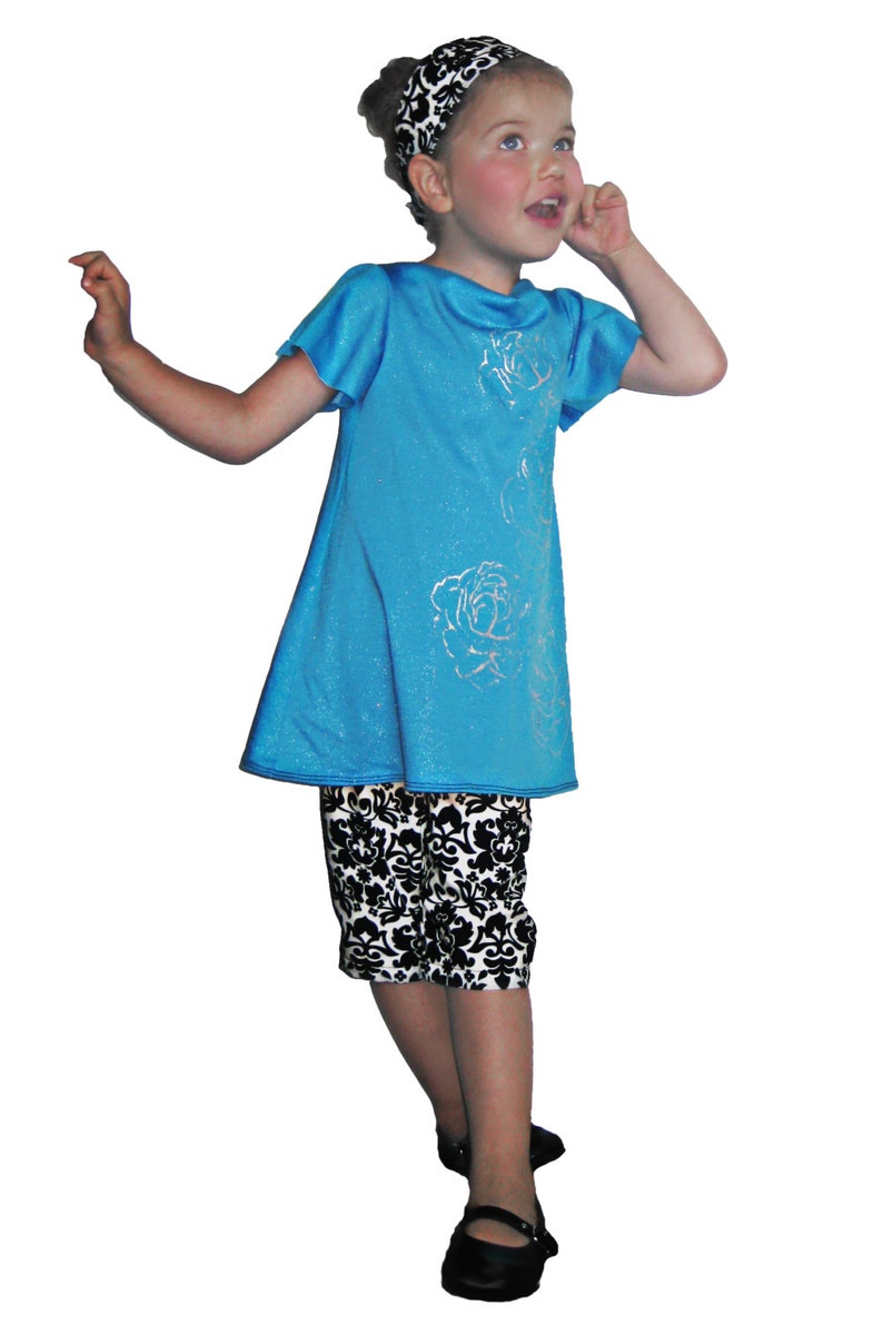 Puede incluir: Una ni&ntilde;a peque&ntilde;a con un vestido turquesa con detalles de rosas plateadas y pantalones capri con estampado en blanco y negro. Tambi&eacute;n lleva una diadema a juego. El conjunto se completa con zapatos negros.