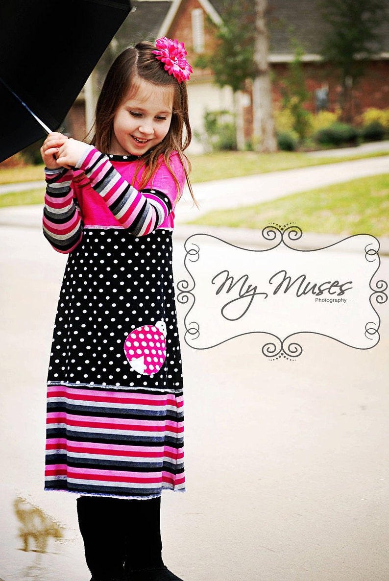 以下が含まれることがあります： ピンク、黒、白の模様のドレスを着た少女。髪には大きなピンクの花が飾られています。ドレスは水玉模様のスカートとストライプの袖が特徴です。黒い傘を持っています。