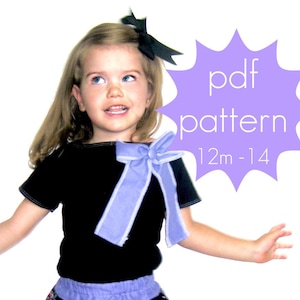 Könnte beinhalten: Ein junges Kind trägt ein schwarzes T-Shirt mit einer hellblauen Schleife und einer schwarzen Schleife im Haar. Das Bild enthält eine lila Stern-Grafik mit dem Text "pdf pattern 12m-14". Das Kind lächelt und blickt nach oben.