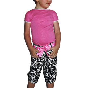 Puede incluir: Conjunto infantil que incluye una camiseta rosa de manga corta con ribete de encaje blanco, combinada con pantalones capri con estampado en blanco y negro. Una diadema a juego y un lazo rosa con lunares adornan la cintura. Zapatos Mary Jane negros completan el conjunto.