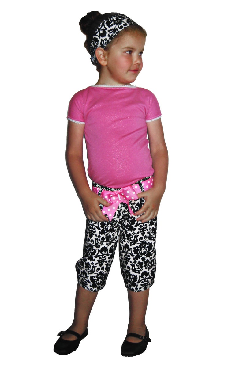 Puede incluir: Conjunto infantil que incluye una camiseta rosa de manga corta con ribete de encaje blanco, combinada con pantalones capri con estampado en blanco y negro. Una diadema a juego y un lazo rosa con lunares adornan la cintura. Zapatos Mary Jane negros completan el conjunto.