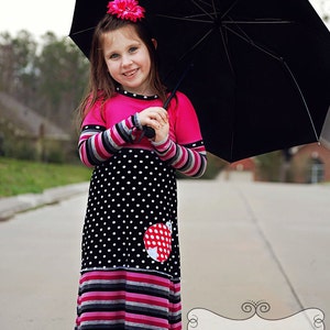 以下が含まれることがあります： 赤いテントウムシのアップリケが付いたピンクと黒の水玉模様のドレスを着た少女が、黒い傘を持っています。ドレスはストライプのアクセントと長袖が特徴です。ピンクの花が彼女の髪を飾っています。
