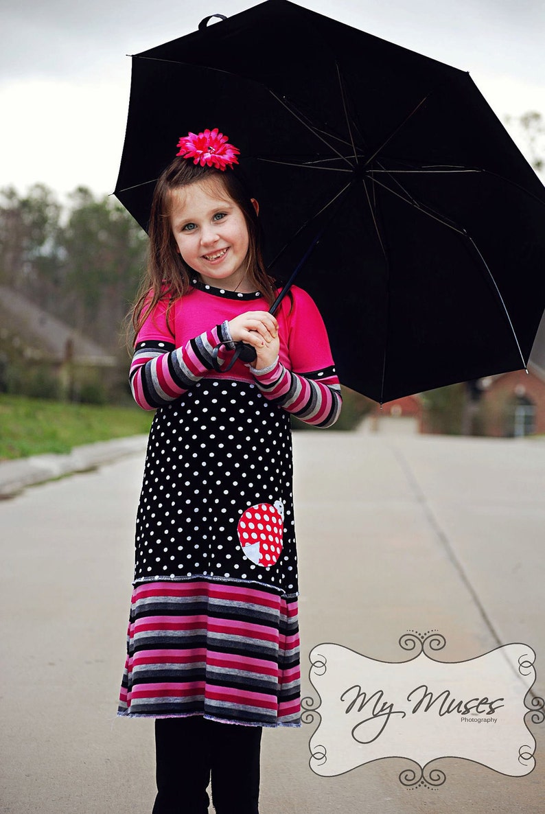 以下が含まれることがあります： 赤いテントウムシのアップリケが付いたピンクと黒の水玉模様のドレスを着た少女が、黒い傘を持っています。ドレスはストライプのアクセントと長袖が特徴です。ピンクの花が彼女の髪を飾っています。