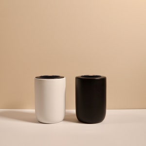 May include: Two cylindrical bathroom toothbrush holders, one white and one black, on a light brown surface.
