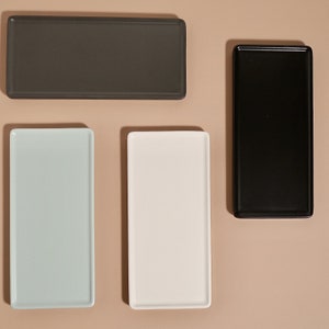May include: Four rectangular serving trays in different colors: gray, light blue, white, and black. The trays are arranged in a row on a light brown surface.