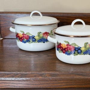 Könnte beinhalten: Set aus drei weißen Emailletöpfen mit Deckeln. Die Töpfe haben einen goldenen Rand und ein farbenfrohes Obst- und Beerenmuster um die Mitte.