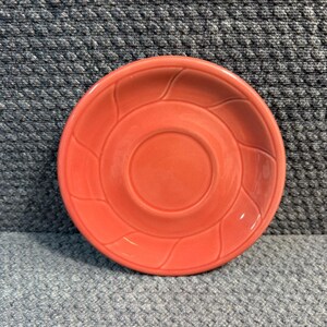 May include: A coral-colored ceramic saucer with a central recessed area and a decorative, raised leaf-like pattern around the rim. The saucer is round and has a glossy finish, suitable for a teacup or small cup.