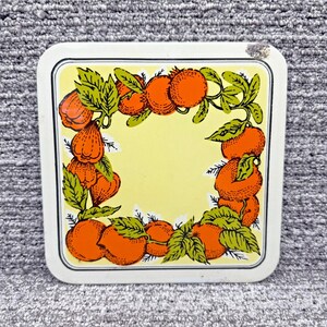 May include: A white square ceramic trivet with a yellow background and a border of orange and green fruit. The fruit is arranged in a circle around the center of the trivet.