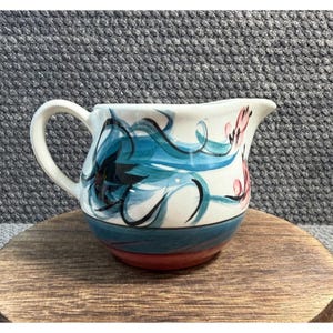 May include: A ceramic creamer pitcher featuring a cream-colored base with hand-painted blue and black floral brushstrokes and pink accents. The pitcher has a curved handle, a pouring spout, and a wide base with a dark blue and red band.