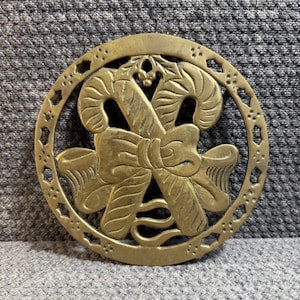 May include: A brass Christmas trivet featuring a candy cane and bow design. The trivet is circular with an openwork border. The candy canes are crossed and tied with a large bow. A sprig of holly is at the top.