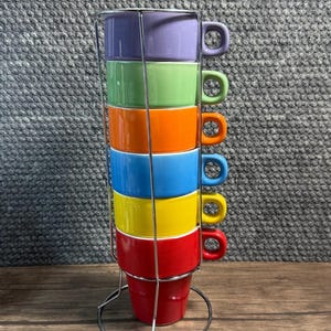 May include: A set of six stacked ceramic mugs in a vertical chrome wire rack. The mugs are arranged in a rainbow color sequence from top to bottom: purple, light green, orange, blue, yellow, and red. Each mug features a small side handle.