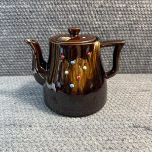 Pode incluir: Uma chaleira de cerâmica castanha vintage com tampa e pega. A chaleira é decorada com pontos vermelhos, brancos e azuis. O bico e a pega são de um castanho mais escuro. A tampa tem um pequeno botão e um padrão decorativo.