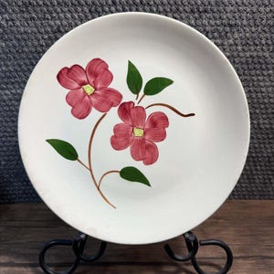 May include: A round white ceramic plate featuring a hand-painted design of two pink flowers with yellow centers, brown stems, and green leaves. The floral motif is centered on the plate, which has a smooth, glossy finish and a simple circular rim.
