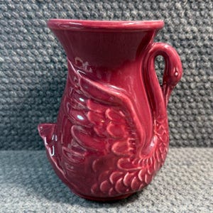 May include: A burgundy ceramic jug with a swan design. The jug has a swan's head as a handle and the body of the swan forms the jug's base. The swan's wings are detailed with textured feathers.