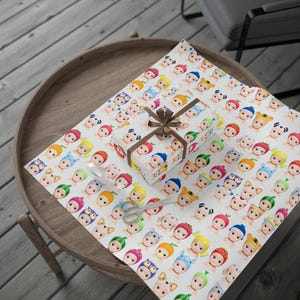 May include: A wrapped gift box and wrapping paper featuring a repeating pattern of colorful baby-like figures with various hats. The box is tied with a brown ribbon. A pair of scissors and a spoon are also on the table.