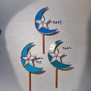May include: Three stained glass crescent moon and star garden stakes in navy, robin egg, and teal. Each stake has a copper-colored stem and is labeled with its color name in handwritten text.
