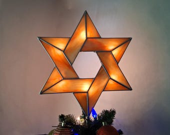 Può includere: Una stella di David in vetro colorato, nei toni dell'arancione e dell'ambra, come decorazione per l'albero. La stella a sei punte è montata su un albero di Natale, con ornamenti e luci visibili. La stella ha un bordo metallico.