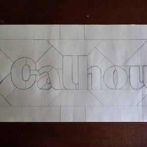 May include: A pencil sketch on paper featuring the name "The Calhouns" in a stylized font. The name is centered within a rectangular frame, with geometric patterns surrounding the text. The design appears to be a template for a sign or artwork.