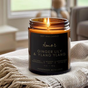 Puede incluir: Un tarro de vela de vidrio marrón con una etiqueta negra que dice "Soy Wax Candle Amor Ginger Lily & Ylang Ylang". La vela está encendida y la etiqueta también incluye "Natural Ingredients", "Wooden Wick", "Handpoured in the UK", "5 oz. 146 g" y "Handmade with Love".