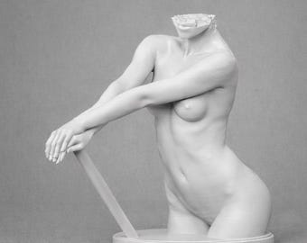 May include: A monochrome sculpture of a female torso, missing the head. The figure is leaning on a rectangular support. The sculpture is smooth and detailed, with visible curves and anatomical features. The background is a solid grey.