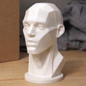 Puede incluir: Busto blanco y geométrico de una cabeza humana. La escultura presenta un diseño angular detallado con rasgos faciales definidos. El busto está montado sobre una base circular. Probablemente sea una pieza de arte decorativa.