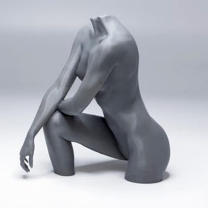 Op de afbeelding: Een grijze sculptuur van een menselijk torso en armen in een zittende pose. De figuur is hoofdloos, met gekruiste armen en gebogen benen. De sculptuur is gemaakt van een glad materiaal en staat tegen een witte achtergrond.
